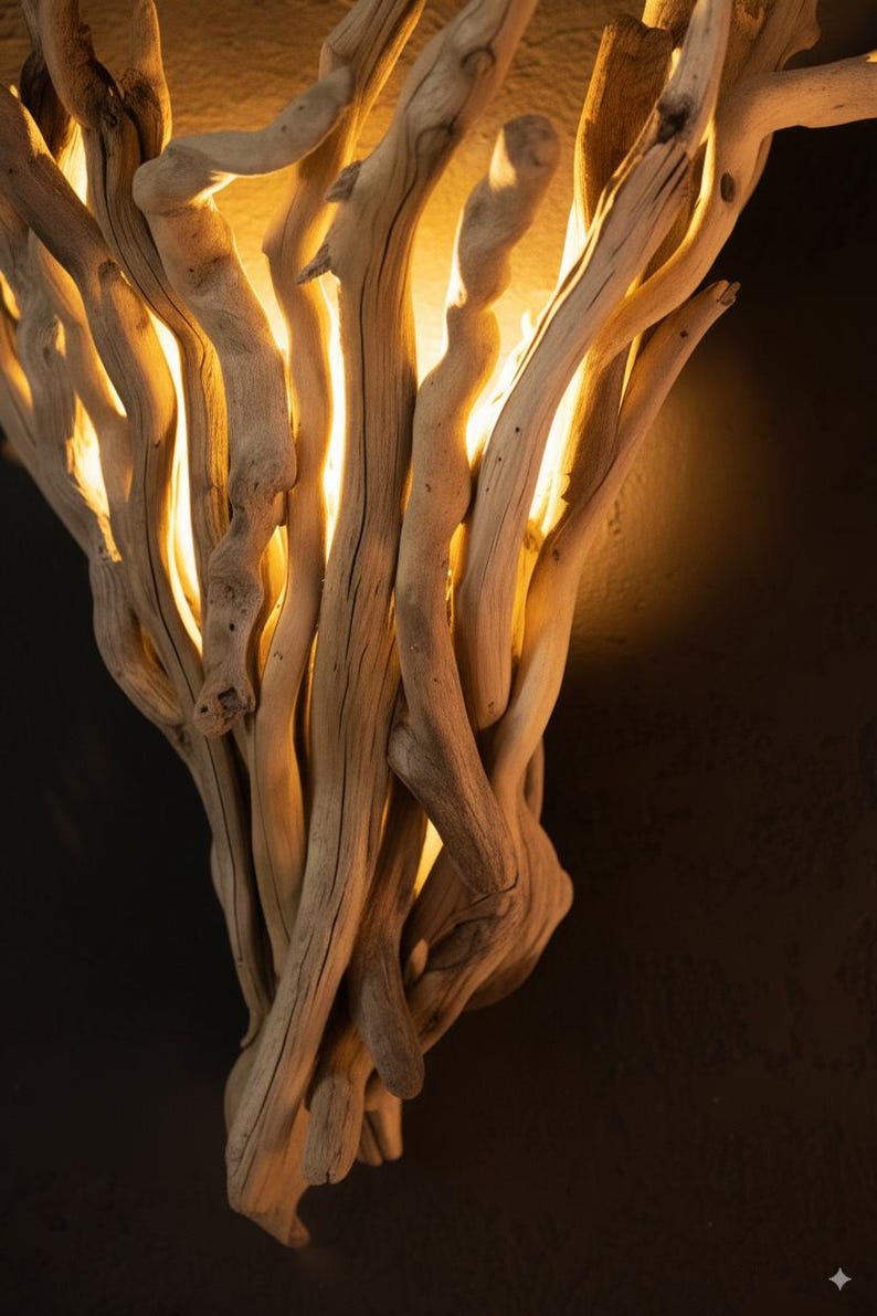 Puede incluir: Un aplique de pared construido con un conjunto de ramas de madera flotante de color claro y aspecto desgastado. La disposici&oacute;n de la madera crea una forma vertical en abanico, con una luz c&aacute;lida y dorada que emana entre las ramas.