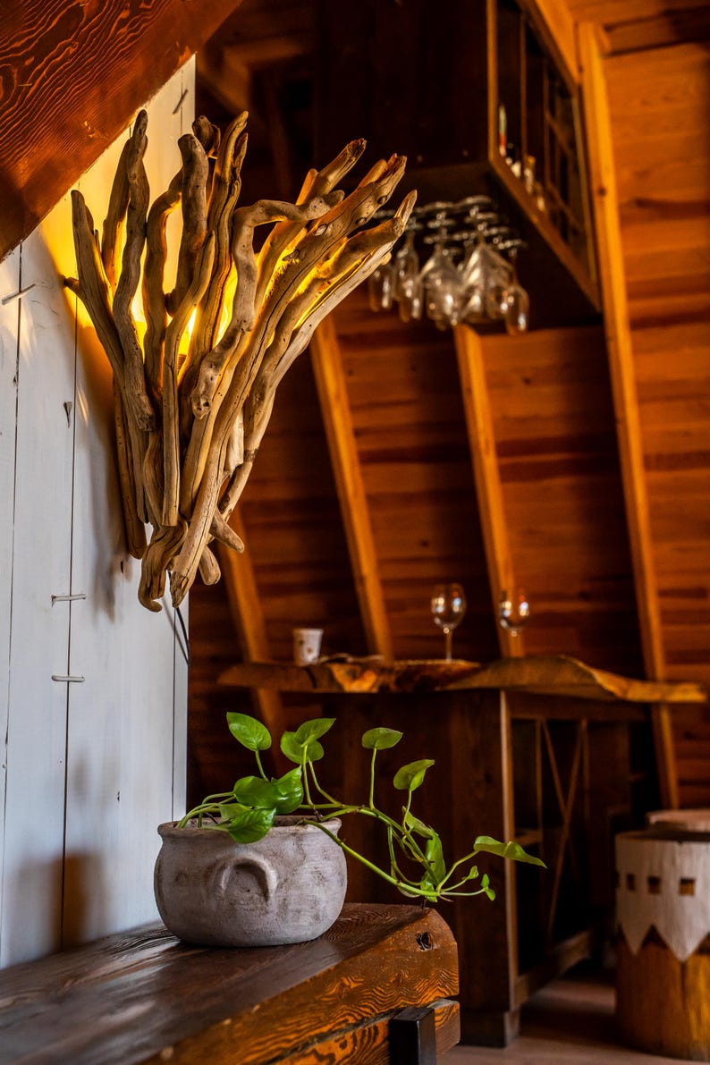 Puede incluir: Un aplique de pared construido con un conjunto de ramas de madera flotante de color marr&oacute;n claro con un brillo c&aacute;lido interno. Debajo, una peque&ntilde;a maceta de cer&aacute;mica gris claro, redonda y con dos asas, contiene una planta verde colgante.