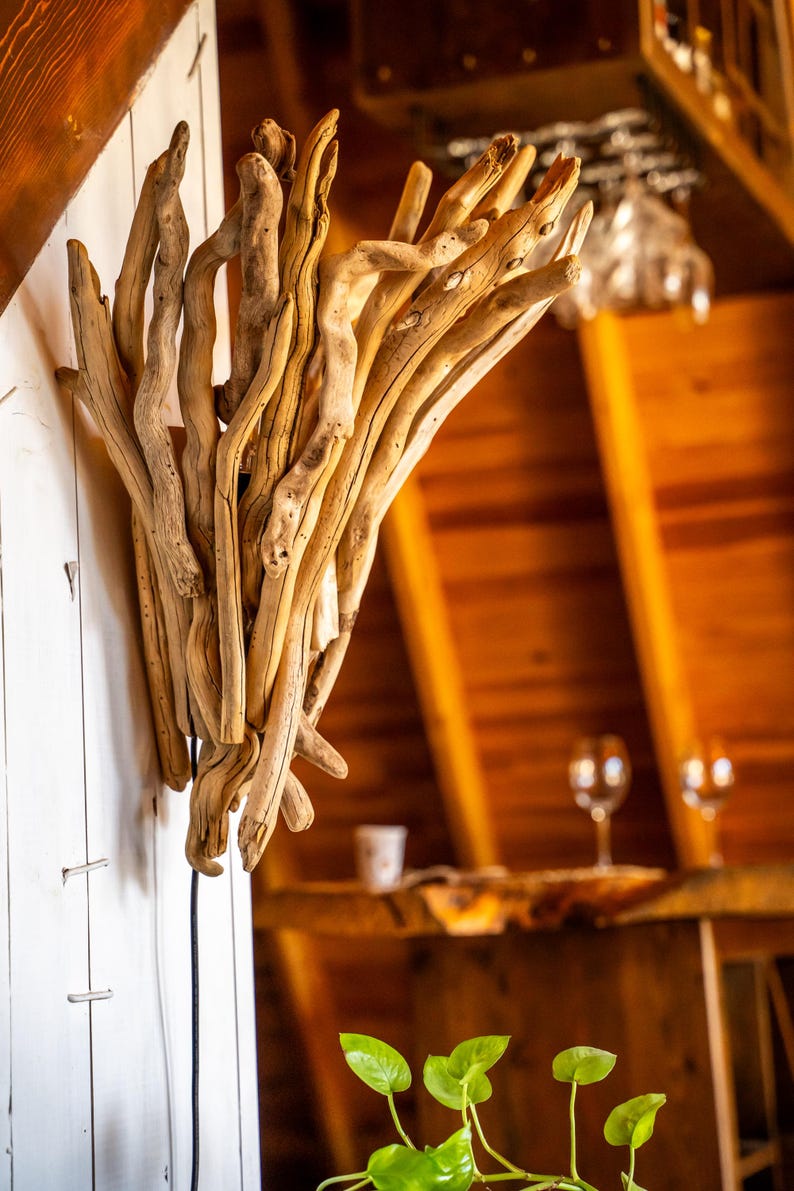 Puede incluir: Un aplique de pared construido a partir de un grupo de ramas de madera flotante de color claro y desgastadas, dispuestas en una formaci&oacute;n vertical en forma de abanico. Las piezas de madera natural var&iacute;an en grosor y textura, con un acabado liso.