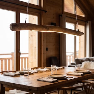 Puede incluir: Una lámpara colgante lineal hecha de una sola pieza de madera flotante natural, suspendida por cables finos sobre una mesa de comedor de madera. La luz cuenta con una tira LED integrada a lo largo de la parte inferior de la madera.