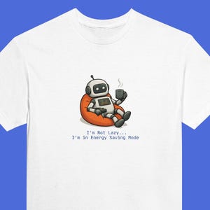 May include: White t-shirt with a cartoon robot on an orange beanbag, holding a coffee mug. The robot has a digital display. Text below reads: "I'm Not Lazy... I'm in Energy Saving Mode." A humorous design.
