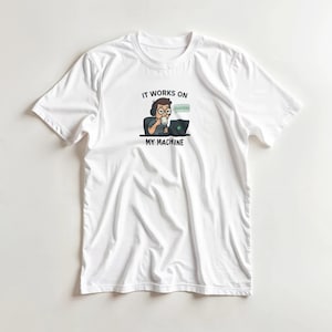 May include: White t-shirt featuring a cartoon graphic of a person working on a laptop and drinking from a mug. The text above the graphic reads "IT WORKS ON MY MACHINE" with a speech bubble that says "SUCCESS".