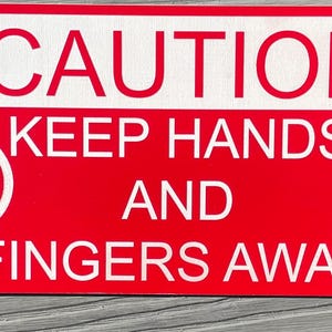 May include: A rectangular red sign with white text and graphics. The top section reads "CAUTION" in large letters. Below, it says "KEEP HANDS AND FINGERS AWAY." White hand symbols with a red slash are on each side.