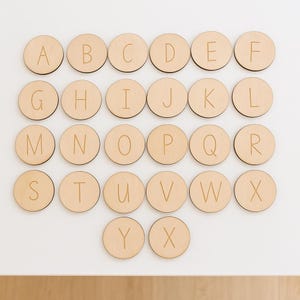 May include: A set of 26 wooden discs, each displaying a letter of the alphabet in a sans-serif font. The discs are arranged in a grid on a white surface. The wood has a natural, light-brown colour. Ideal for educational purposes.