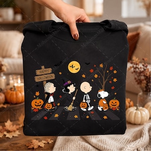 May include: A black sweatshirt featuring a graphic of Peanuts characters trick-or-treating. The design includes a wooden sign reading Happy Halloween, a full moon, bats, autumn leaves, and characters dressed as a vampire, witch, skeleton, and ghost.