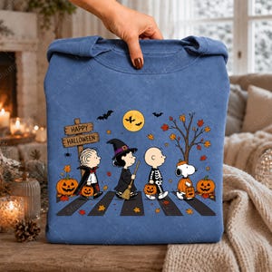 May include: A blue crewneck sweatshirt featuring a graphic of Peanuts characters dressed in Halloween costumes walking across a crosswalk. The design includes a wooden sign reading Happy Halloween, a yellow moon, bats, autumn leaves, and pumpkins.