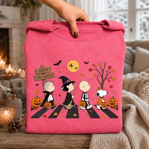 May include: A bright pink sweatshirt featuring a graphic of Peanuts characters dressed in Halloween costumes walking across a crosswalk. The design includes a wooden sign that reads Happy Halloween, pumpkins, bats, a full moon, and autumn leaves.