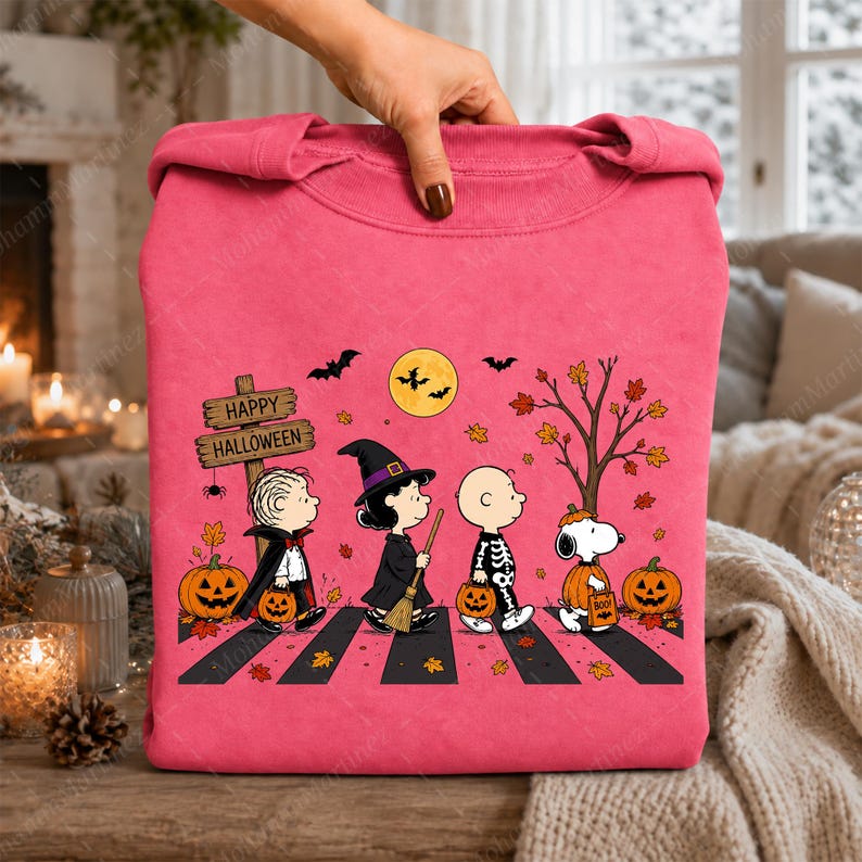 May include: A bright pink sweatshirt featuring a graphic of Peanuts characters dressed in Halloween costumes walking across a crosswalk. The design includes a wooden sign that reads Happy Halloween, pumpkins, bats, a full moon, and autumn leaves.