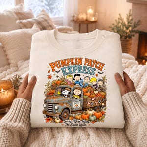 May include: A cream-colored crewneck sweatshirt featuring a graphic of a vintage blue truck filled with pumpkins. The text reads PUMPKIN PATCH EXPRESS and Sorry... We're Taking The Scenic Route. Characters from Peanuts are shown in the truck bed.