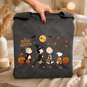 May include: A charcoal gray crewneck sweatshirt featuring a graphic of four cartoon characters dressed in Halloween costumes. The design includes a wooden sign reading Happy Halloween, a yellow moon, bats, orange pumpkins, and autumn leaves.