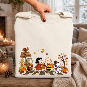 May include: A cream-coloured crewneck sweatshirt featuring a Peanuts Thanksgiving graphic. The design shows characters walking with autumn harvest items, a turkey on a sign that reads Happy Thanksgiving, and falling leaves in orange and red.