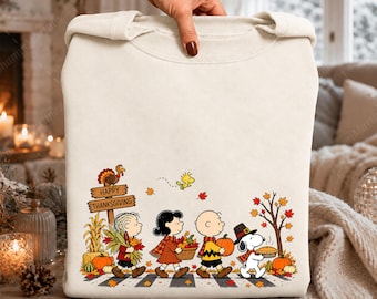 May include: A cream-colored crewneck sweatshirt featuring a Peanuts Thanksgiving graphic. The design shows characters walking with autumn harvest items, a turkey on a sign that reads Happy Thanksgiving, and falling leaves in orange and red.