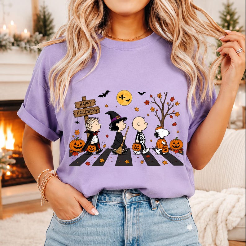May include: A light purple t-shirt featuring a graphic of Peanuts characters dressed in Halloween costumes walking across a crosswalk. The design includes a wooden sign that reads Happy Halloween, orange pumpkins, autumn leaves, and bats.