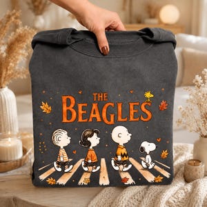 Könnte beinhalten: Ein anthrazitfarbenes Sweatshirt mit Rundhalsausschnitt und einer Grafik von vier Zeichentrickfiguren, die über einen Zebrastreifen gehen. Der Text "THE BEAGLES" steht in Orange darüber, umgeben von kleinen Herzen und Herbstblättern.