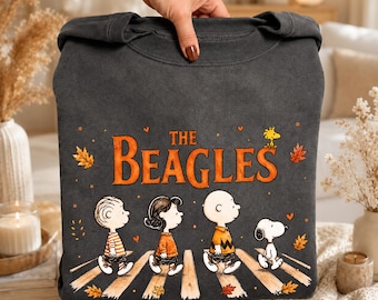 May include: A charcoal gray crewneck sweatshirt featuring a graphic of four cartoon characters walking across a crosswalk. The text "THE BEAGLES" is printed in orange above the characters, surrounded by small orange hearts and autumn leaves.