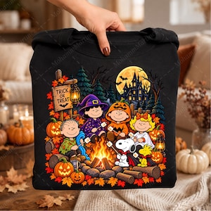 May include: A black sweatshirt featuring a graphic of Peanuts characters sitting around a campfire at night. The scene includes a wooden sign that reads Trick or Treat, a haunted house, pumpkins, autumn leaves, and a lantern in a forest setting.