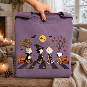 May include: A purple crewneck sweatshirt featuring a graphic of Peanuts characters dressed in Halloween costumes walking on a crosswalk. The design includes a wooden sign reading Happy Halloween, pumpkins, bats, a full moon, and autumn leaves.