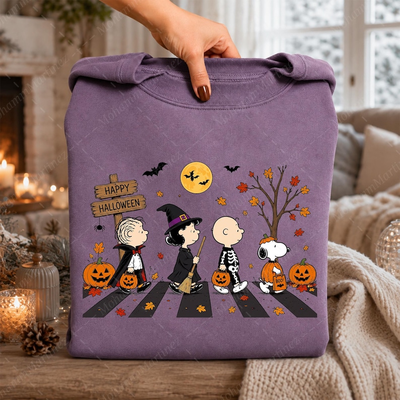 May include: A purple crewneck sweatshirt featuring a graphic of Peanuts characters dressed in Halloween costumes walking on a crosswalk. The design includes a wooden sign reading Happy Halloween, pumpkins, bats, a full moon, and autumn leaves.