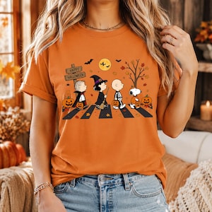 May include: An orange short-sleeve t-shirt featuring a graphic of Peanuts characters dressed in Halloween costumes walking across a crosswalk. The design includes a sign that reads Happy Halloween, pumpkins, a full moon, and autumn leaves.