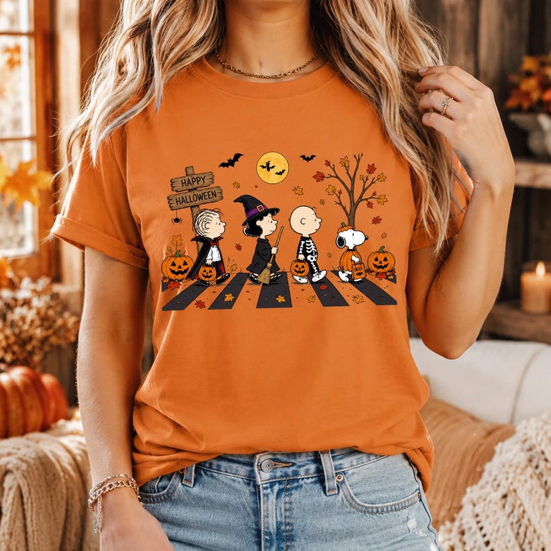 May include: An orange short-sleeve t-shirt featuring a graphic of Peanuts characters dressed in Halloween costumes walking across a crosswalk. The design includes a sign that reads Happy Halloween, pumpkins, a full moon, and autumn leaves.