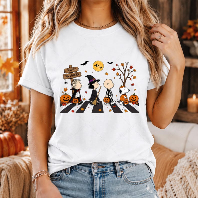 May include: A white t-shirt featuring a graphic of Peanuts characters dressed in Halloween costumes walking across a crosswalk. The design includes a wooden sign reading Happy Halloween, orange pumpkins, a yellow moon, bats, and autumn leaves.
