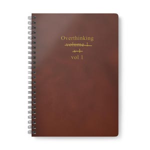 May include: A brown spiral-bound notebook with the words "Overthinking" crossed out and replaced with "vol 1" in gold lettering. The notebook has a leather-like texture.