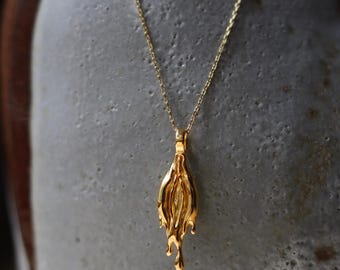 Könnte beinhalten: Eine goldfarbene Anhängerhalskette mit einem einzigartigen, abstrakten Design. Der Anhänger hat eine strukturierte, organische Form mit einem tropfenden Effekt am unteren Ende. Die Halskette ist zierlich und goldfarben.