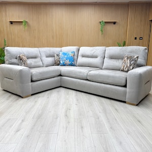 May include: A large, light grey corner sofa with three back cushions and two armrests. Decorative cushions in blue and patterned designs add colour. The sofa is on a light wood floor. The sofa is a modern design.