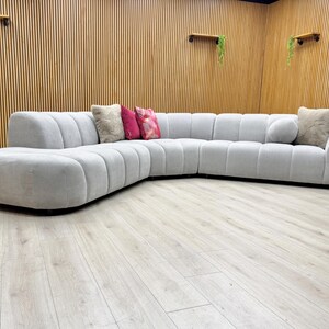 May include: A large, light grey sectional sofa with a rounded design. The sofa has a series of vertical, padded sections and is accompanied by several decorative cushions in shades of pink and beige. The sofa is placed on a light wood floor.