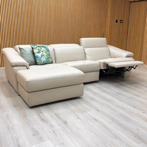 May include: A cream leather sectional sofa with a chaise longue and reclining function. The sofa has a contemporary design. Two decorative cushions with nature-themed patterns add colour. The sofa is set against a wood-panelled wall.