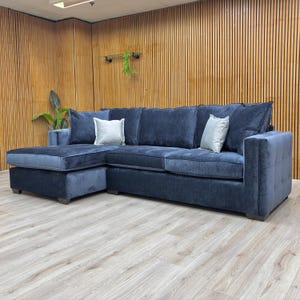 May include: A large, navy blue corner sofa with a chaise longue on the left. The sofa features plush cushions and two silver scatter cushions. The armrests have a quilted design. The sofa sits on a light wood floor.