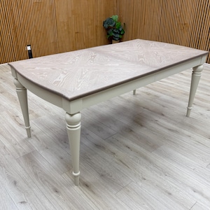 May include: A rectangular dining table with a light wood grain top and a dark brown trim. The table has four cream-coloured, turned legs. The table is set on a light wood floor.