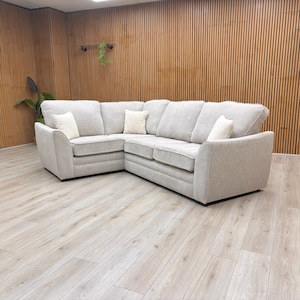May include: A large, light grey corner sofa. The sofa has a textured fabric, with white accent cushions. The room has light wood flooring and a wood-panelled wall.