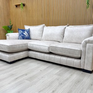 May include: A beige corner sofa with a chaise lounge. The sofa features three seat cushions and a blue decorative cushion with a white bird design. The sofa is set on a light wood floor.