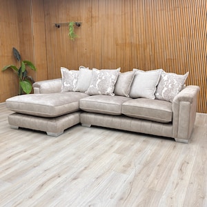 May include: A beige corner sofa with a chaise longue on the left. It features several decorative cushions with floral patterns. The sofa has a quilted design on the cushions and armrests. The sofa sits on silver feet.