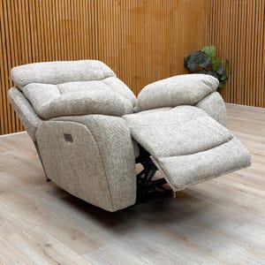 May include: A light grey recliner chair with a textured fabric. The chair is partially reclined, with the footrest extended. The headrest and armrests are well-padded. The chair sits on a black metal frame. The background features wooden paneling.