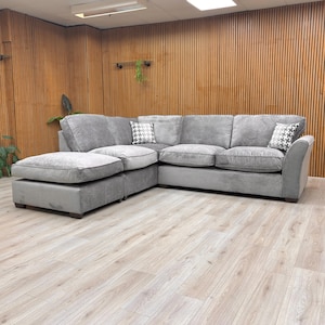 May include: A large, grey corner sofa with a chaise longue on the left. The sofa features soft cushions and two patterned scatter cushions. The room has wooden paneling and light-coloured flooring. A comfortable seating option.