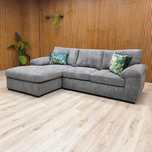 May include: A grey corduroy corner sofa with a chaise longue on the left. Two decorative cushions with floral designs are on the sofa. The room has wooden paneling and a light wood floor.