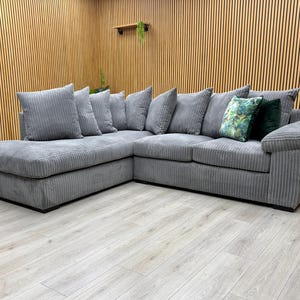 May include: A grey corduroy corner sofa with a chaise longue, featuring several matching cushions. The sofa is set against a wooden panelled wall. Two decorative cushions have a green floral pattern. The sofa is approximately 250 cm long.