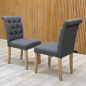 May include: Two grey upholstered dining chairs with button detailing and curved backs. The chairs have light-coloured wooden legs. The chairs are set against a wooden slatted wall. Each chair is designed for comfort and style.
