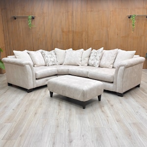 May include: A beige corner sofa with a matching footstool. The sofa has several white and patterned cushions. The furniture is set on a light wood floor, with a wooden slatted wall in the background.