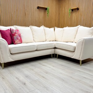 May include: Cream-coloured corner sofa with a curved design. The sofa has multiple cushions and two decorative pillows in shades of pink. The sofa sits on gold-coloured legs. The background features wooden panelling.