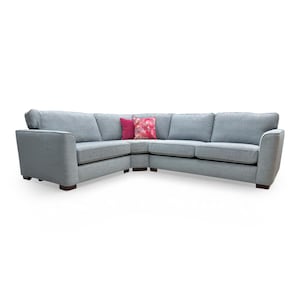 May include: A light blue corner sofa with a curved design. The sofa features a soft, textured fabric and dark wooden feet. Two decorative cushions, one magenta and one floral, provide a contrast to the neutral sofa. The sofa measures approximately 250 cm.