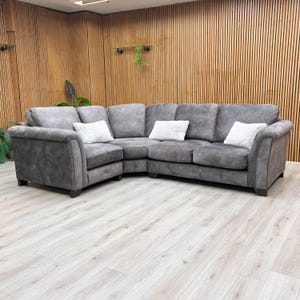 May include: A large, grey corner sofa with three white cushions. The sofa has a curved design and sits on dark wooden legs. The background features wooden paneling and a light-coloured floor.