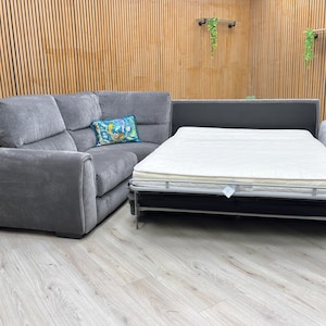 May include: A grey corner sofa with a pull-out bed. The sofa has a soft, textured look and a patterned cushion. The bed is extended, showing a white mattress. The sofa is on a light wood floor, with a wooden panelled wall behind.