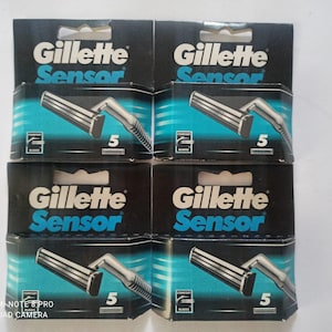 Könnte beinhalten: Mehrere Schachteln mit Gillette Sensor Rasierklingen sind gestapelt. Jede Schachtel ist schwarz mit dem Gillette-Logo in Weiß und Blau. Das Wort "Sensor" ist blau. Die Schachteln zeigen eine silberne Rasierklinge und die Zahl "5".