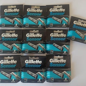 May include: A selection of Gillette Sensor razor blade cartridges in their original packaging. The black packaging has teal accents and features the brand name and product details. Each pack contains five blades.
