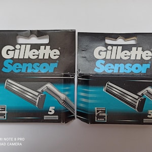 Könnte beinhalten: Zwei Packungen Gillette Sensor Rasierklingen. Die Verpackung ist schwarz mit dem Marken- und Produktnamen in Weiß und Blau. Jede Packung zeigt einen silbernen Rasierer und die Zahl "5". Auf der Unterseite der Verpackung steht "COMFORT BLADES".