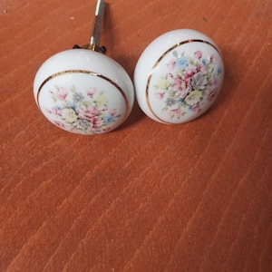 Pode incluir: Dois puxadores de armário de cerâmica branca com desenhos florais em rosa, azul e amarelo. Cada puxador tem uma faixa dourada e uma haste de metal para fixação. Os puxadores são provavelmente para móveis.