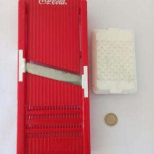 May include: A red Coca-Cola branded mandoline slicer with a stainless steel blade and a white food holder. The slicer has a ridged design and a rectangular shape. A coin is next to the slicer.